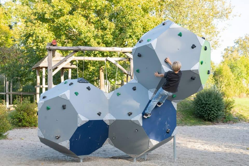 Ein Kind klettert an einem mehrteiligen Klettergerät mit grauen und blauen Flächen sowie grünen und schwarzen Griffen.